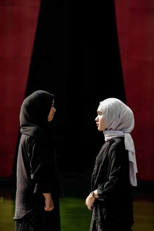 Free Two women wearing hijabs stand face to face against a black and red background. Stock Photo