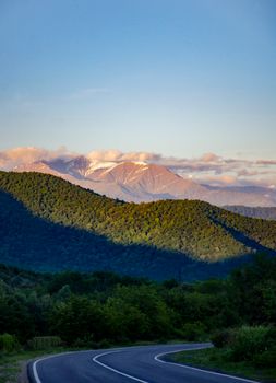 A winding road through verdant hills with sunlit snow-capped mountains and a bright sky.