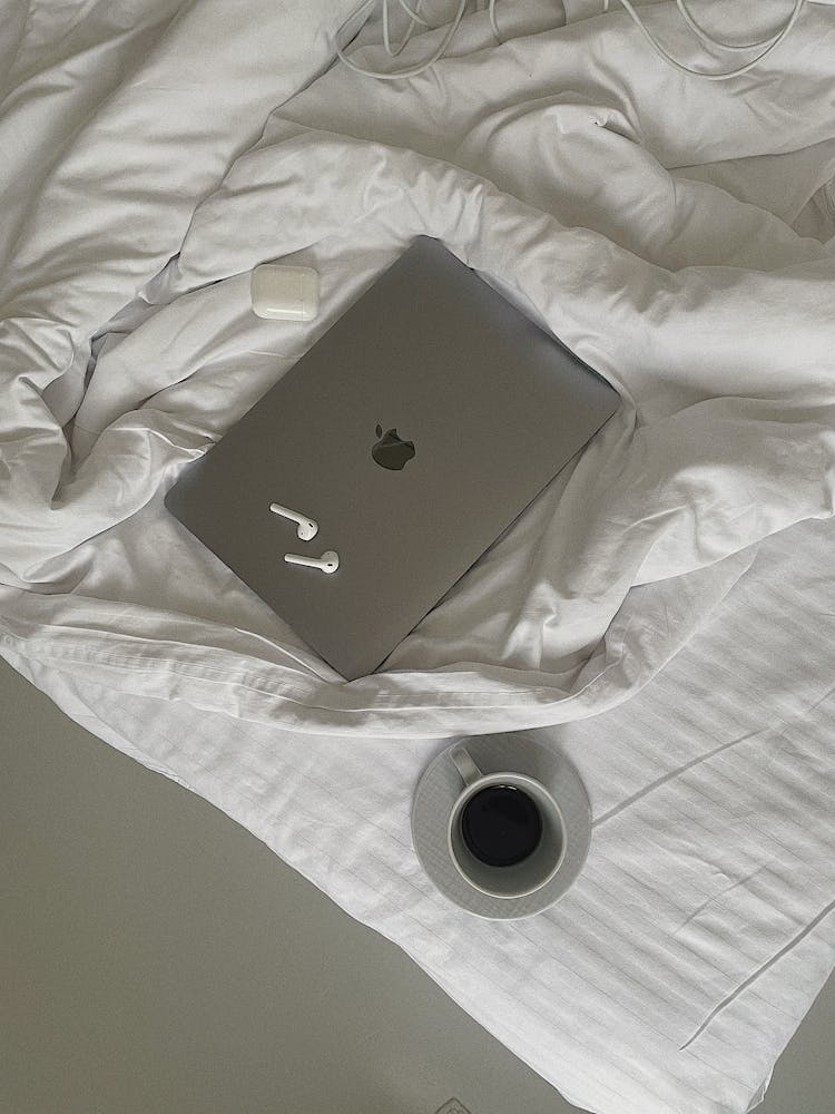 A Laptop And Airpods On The Bed Near The Cup Of Coffee