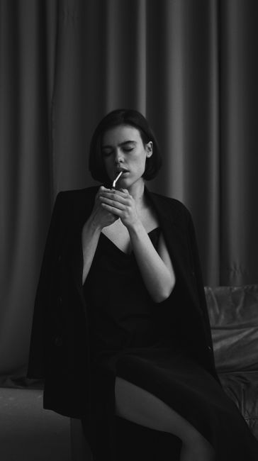 A woman in a black dress and coat smokes a cigarette, creating a moody and elegant atmosphere.