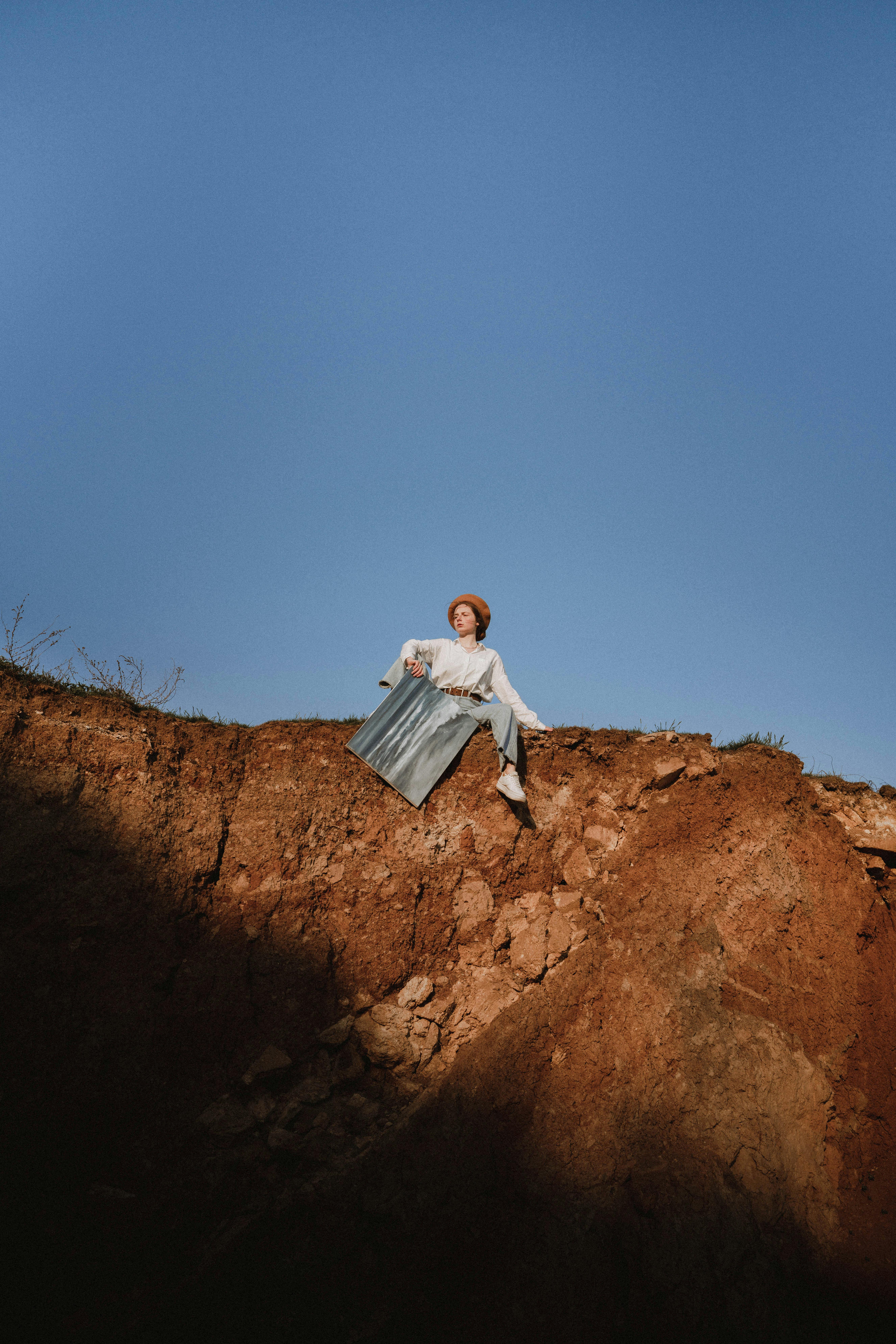 A woman in a hat sits on a rocky cliff edge under a vivid blue sky, embracing nature.