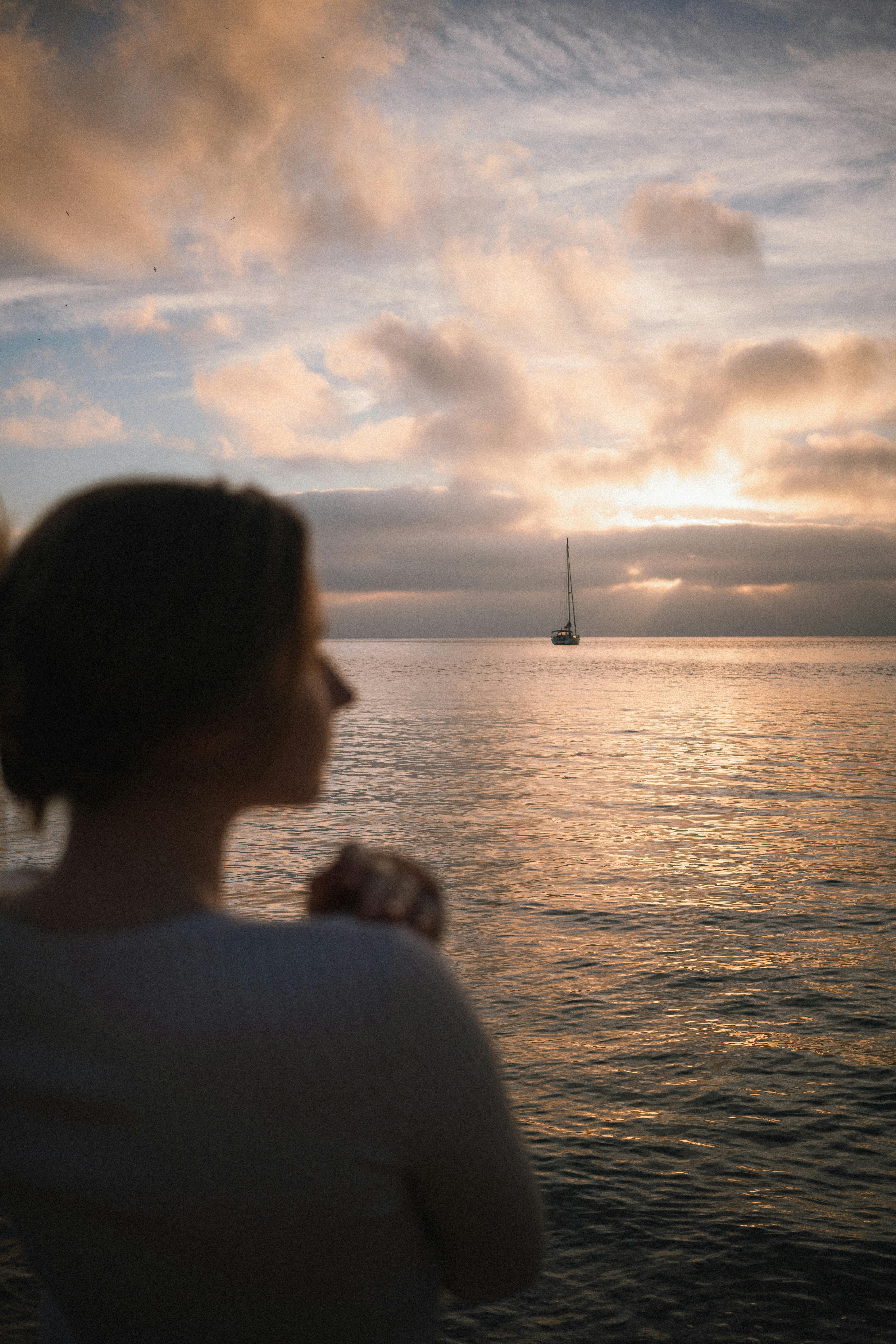 A calming sunset view across the ocean with a distant sailboat and a blurred silhouette in the foreground.