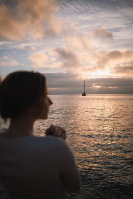 A calming sunset view across the ocean with a distant sailboat and a blurred silhouette in the foreground.