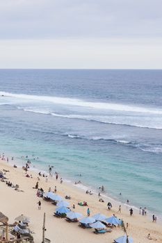 A vibrant beach in Bali, Indonesia with tourists enjoying under blue umbrellas.