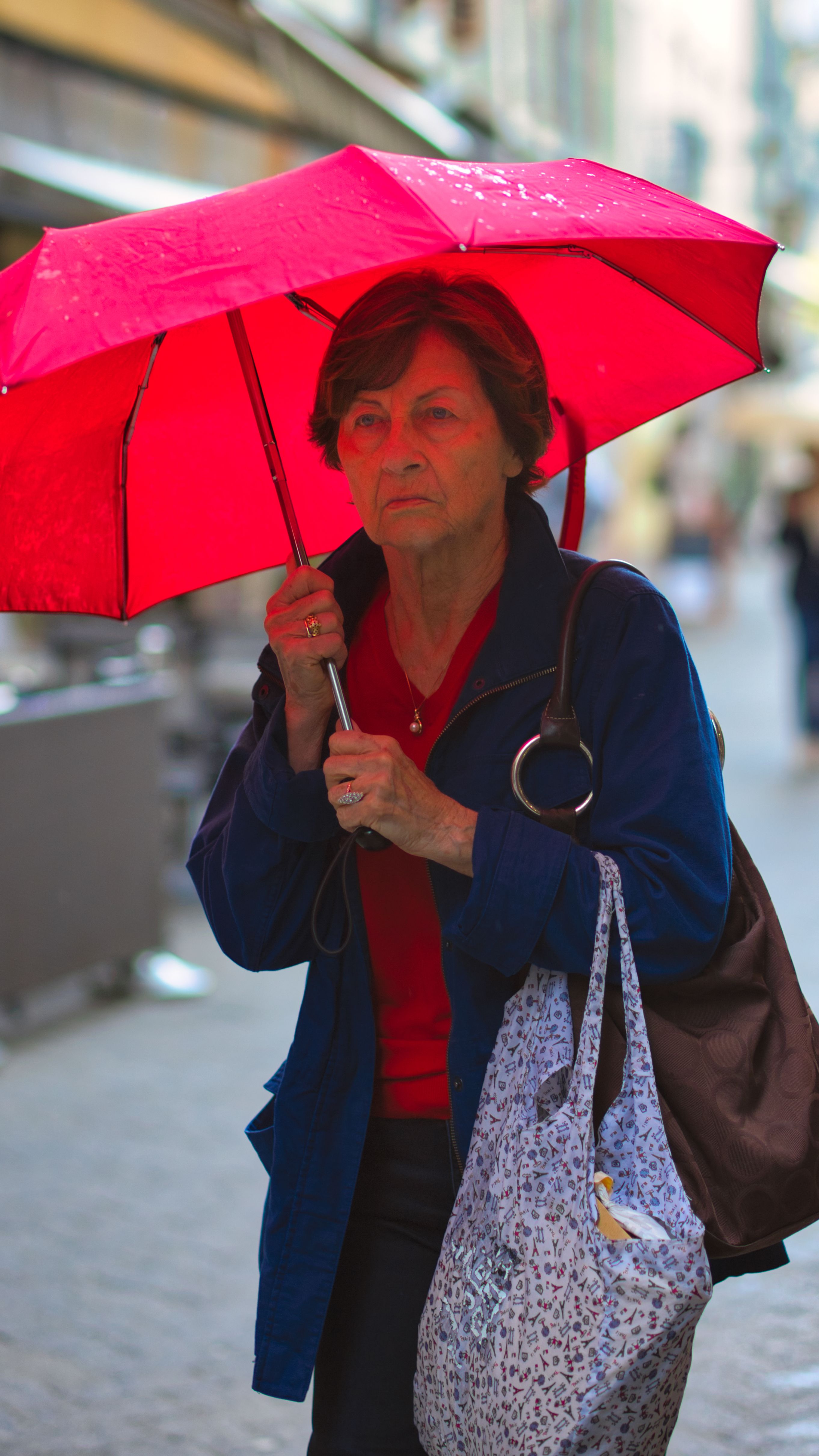 A senior woman carrying a red umbrella walks on a city street captured on a rainy day.