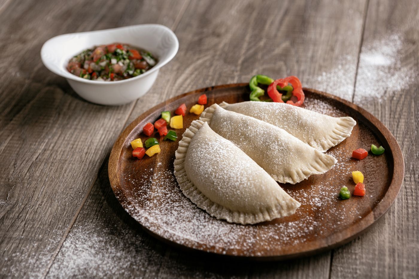Three empanadas on a wooden plate with powdered sugar, served with dipping sauce on a wooden table.