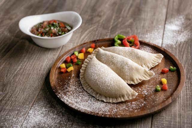 Three empanadas on a wooden plate with powdered sugar, served with dipping sauce on a wooden table.