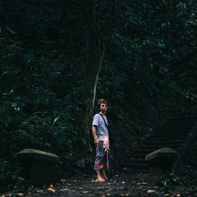 A person in colorful attire standing on a serene forest pathway surrounded by lush greenery.