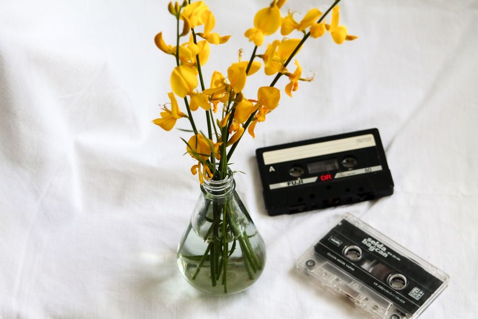 Still life scene with vintage cassette tapes and yellow flowers in a glass vase on white background.