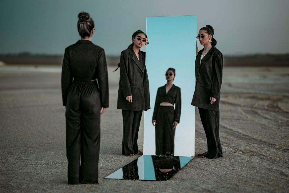 Elegant woman in black clothing reflects in a mirror placed outdoors, creating a surreal fashion scene.