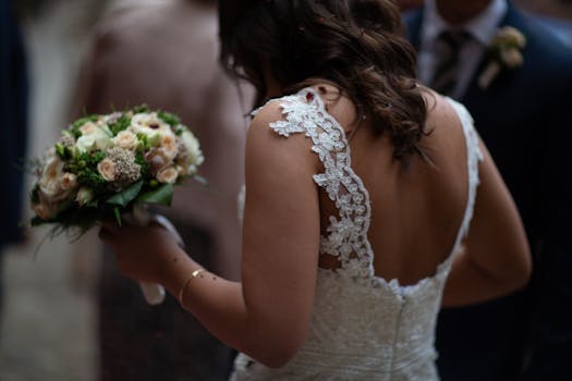A bride in a lace gown holds a beautiful bouquet, viewed from behind at a wedding ceremony.