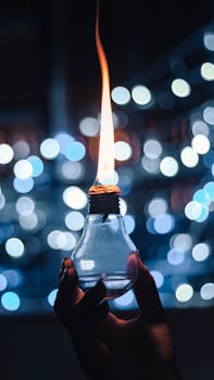A hand holds a light bulb with a unique flame effect against a glowing bokeh backdrop.