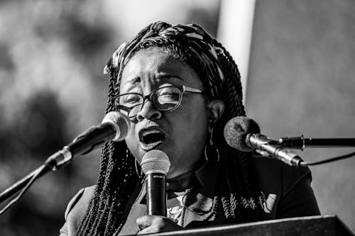 A Grayscale Photo of a Woman Talking in Front of the Microphones