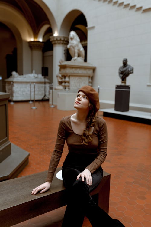 Photo of a Woman in a Brown Beret Sitting with Crossing Legs on a Wooden Bench in a Museum