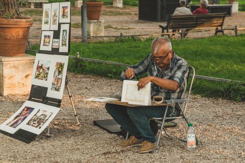 Free An elderly artist focused on sketching in a tranquil outdoor park setting, surrounded by his artwork. Stock Photo