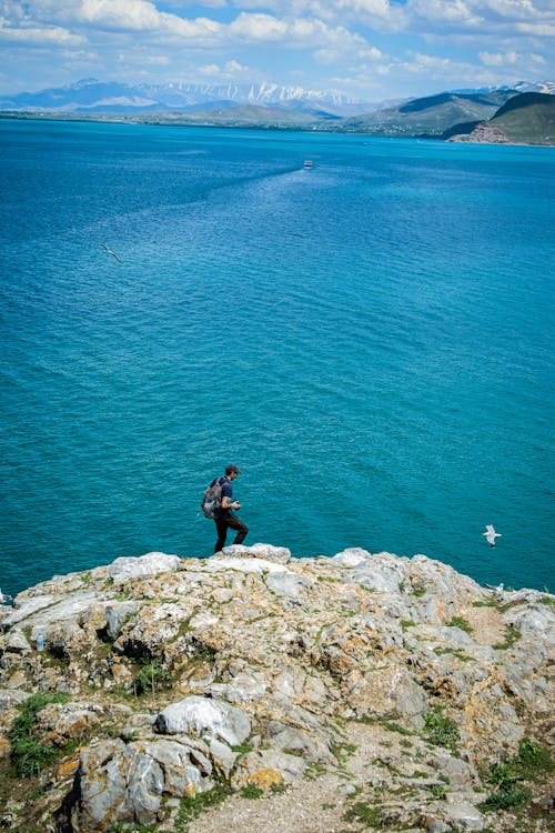 Man With Backpack Walking on Cliff Near Blue Sea