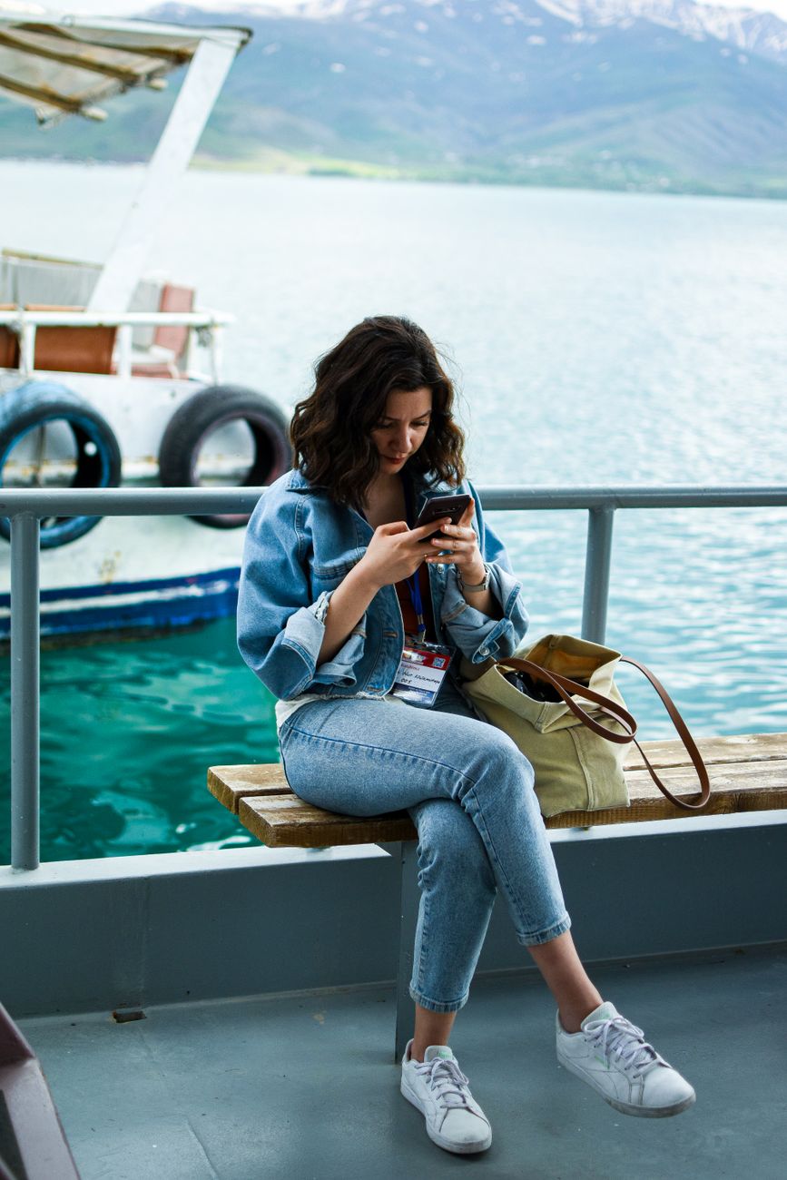 Woman sitting on ferry bench with phone, relaxed atmosphere. Background of serene water and distant mountains.