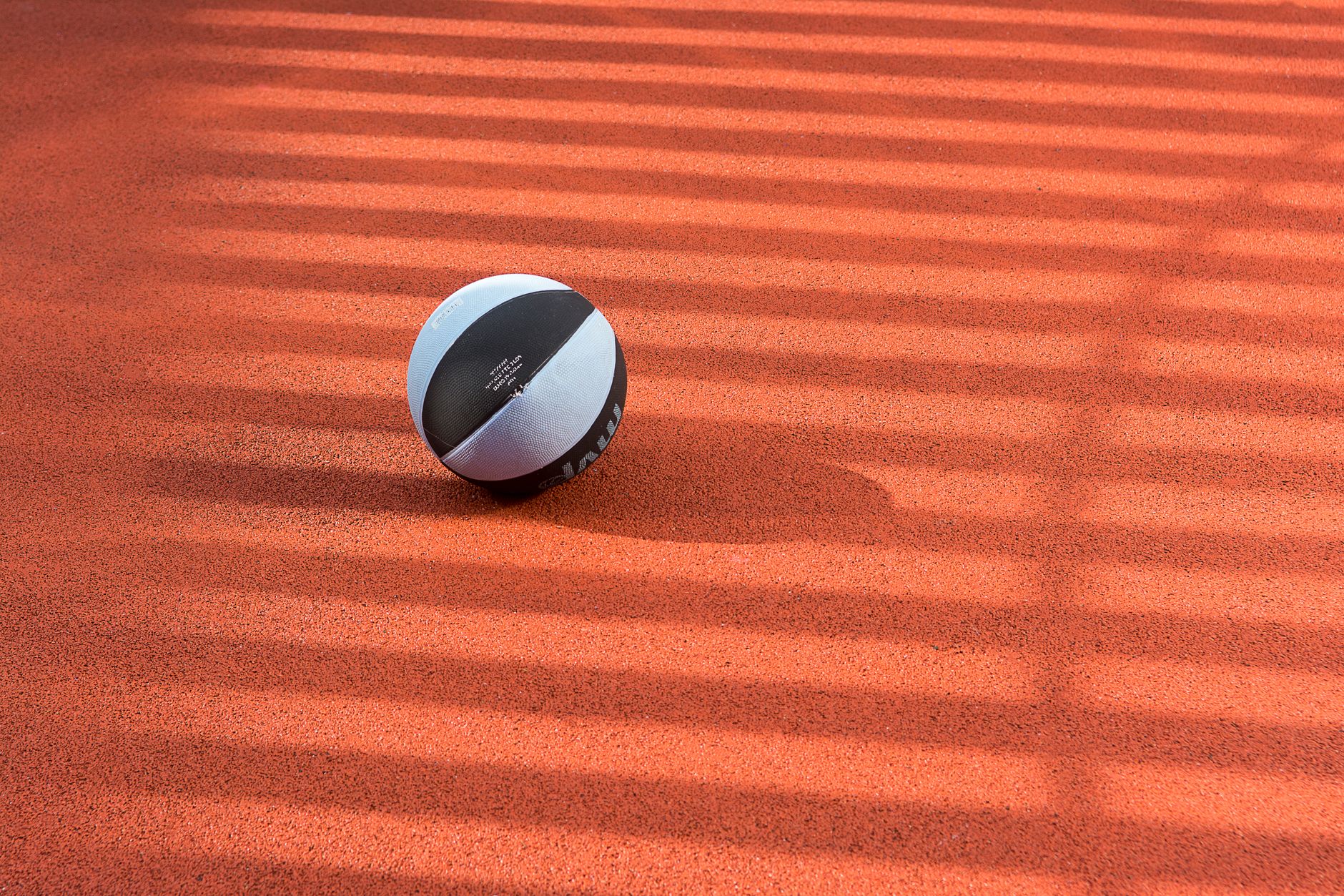 A black and white basketball sits on a sunlit court with shadow stripes, creating an artistic effect.