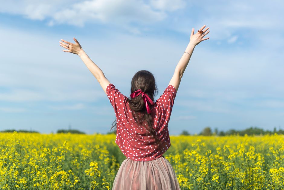 A woman in a polka dot shirt raising her arms in a bright yellow flower field, expressing freedom and joy