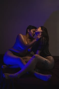 Two women embracing in a sensual scene under moody blue lighting.