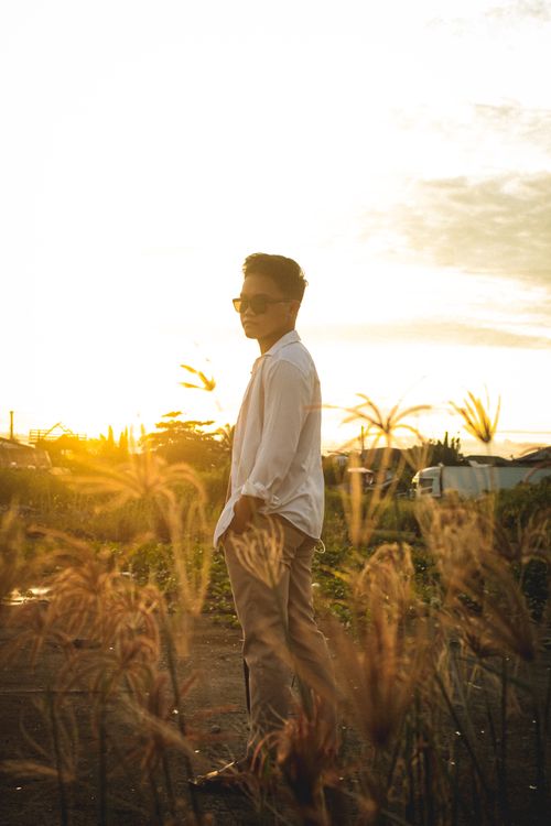 Free A fashionable man standing in a rural field at sunset, wearing sunglasses and a white long sleeve shirt. Stock Photo