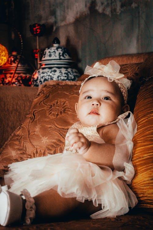 Free Baby in White Dress Lying on Brown Floral Sofa Stock Photo