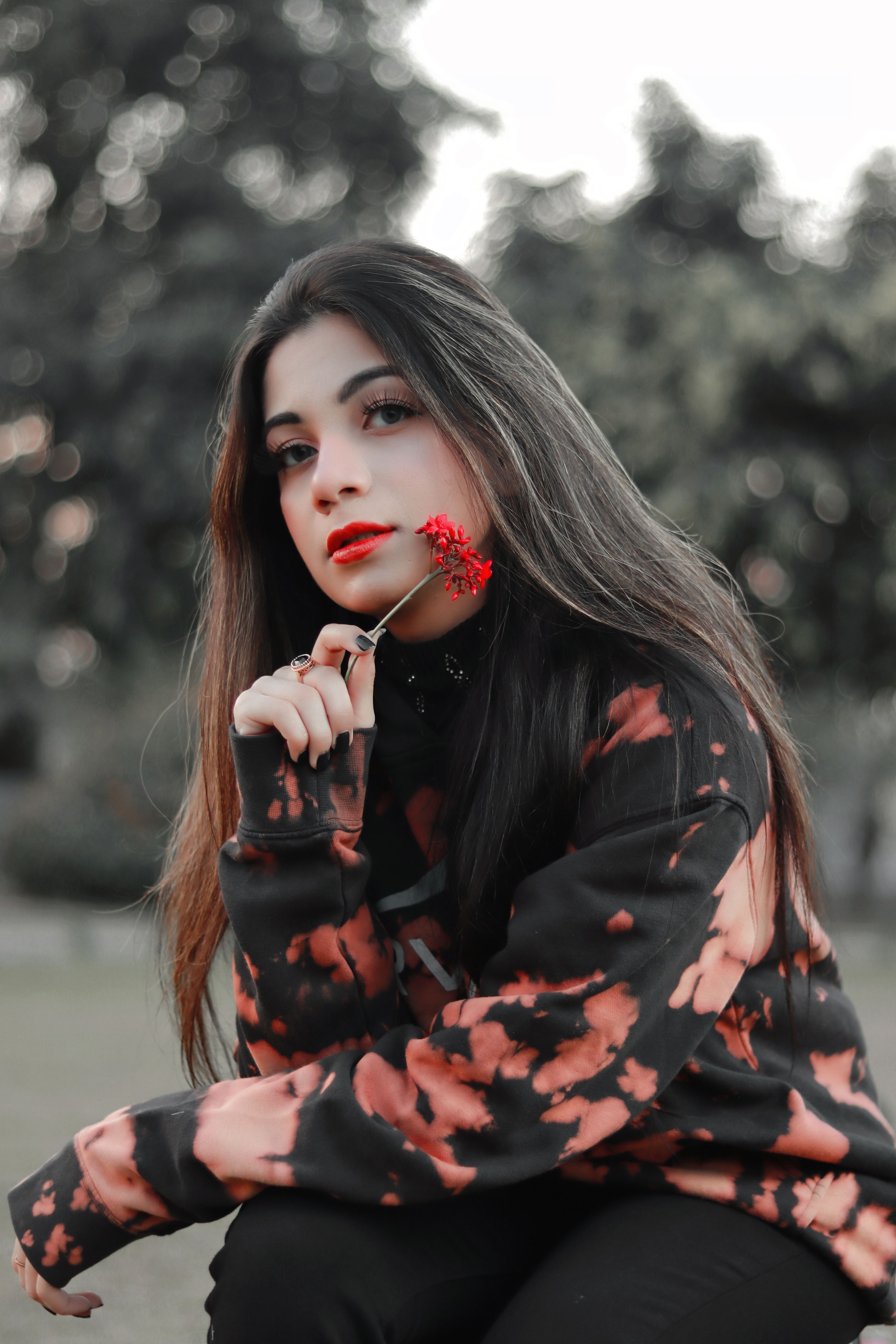 A young woman in a stylish jacket holding a red flower, set against a blurred natural background.