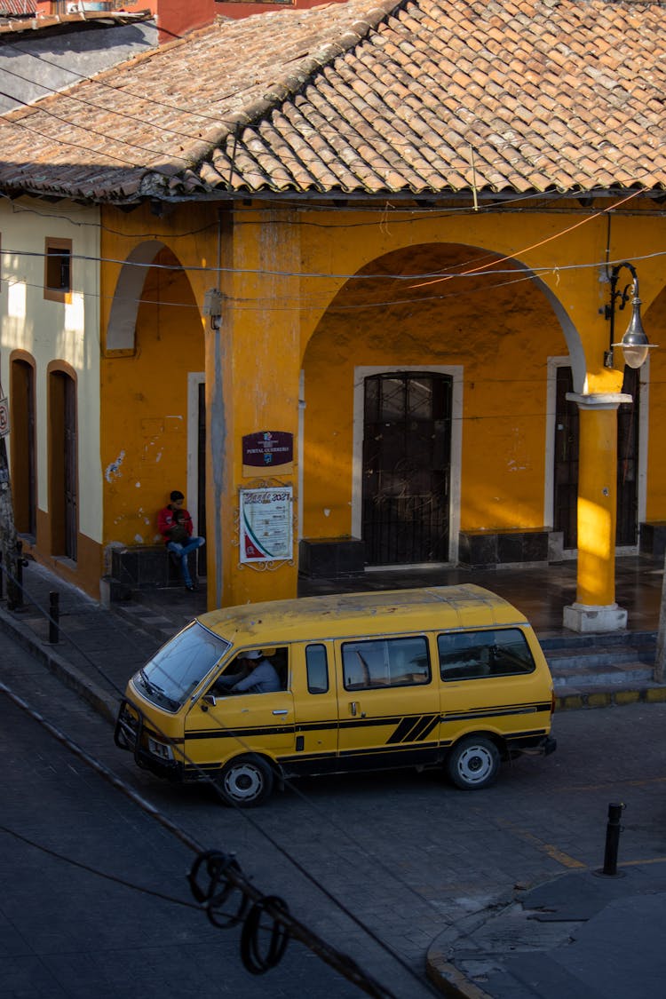 A  Person Driving An Old Yellow Van Near A Yellow Building