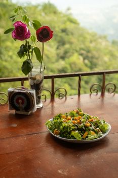 Fresh vegetable salad with colorful roses on a wooden table set in lush greenery outdoors.