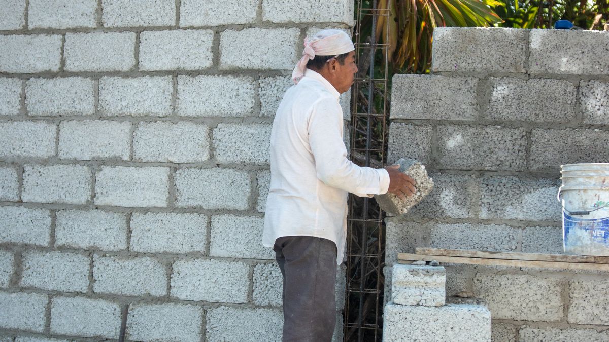 Pedreiro de camisa branca e lenço na cabeça levantando uma parede de blocos de concreto