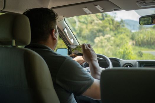 A man uses a smartphone to capture a scene outside while driving in a car interior during the day.