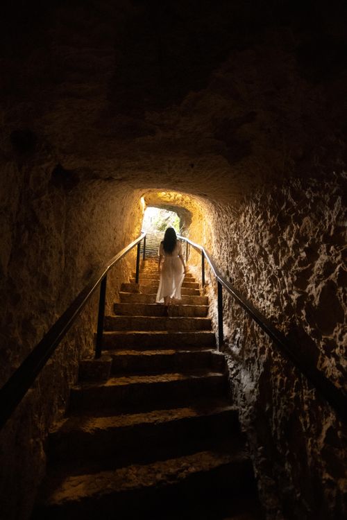 Free A solitary woman walks up stone stairs in an ancient cave tunnel, lit warmly. Stock Photo