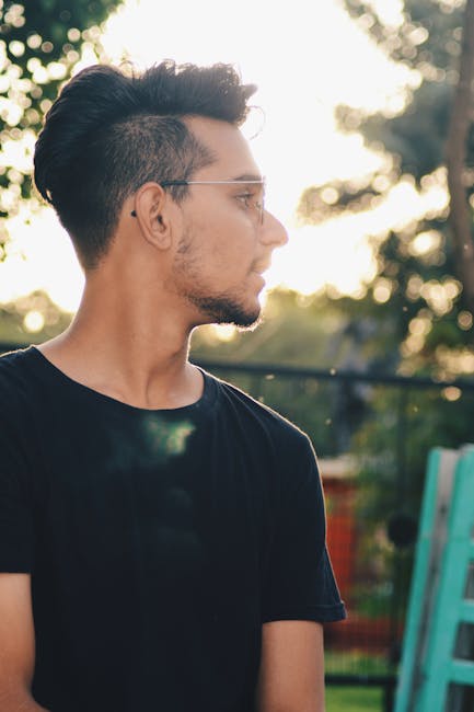 A stylish man in a black t-shirt viewed in profile outdoors during sunset, showcasing casual fashion.