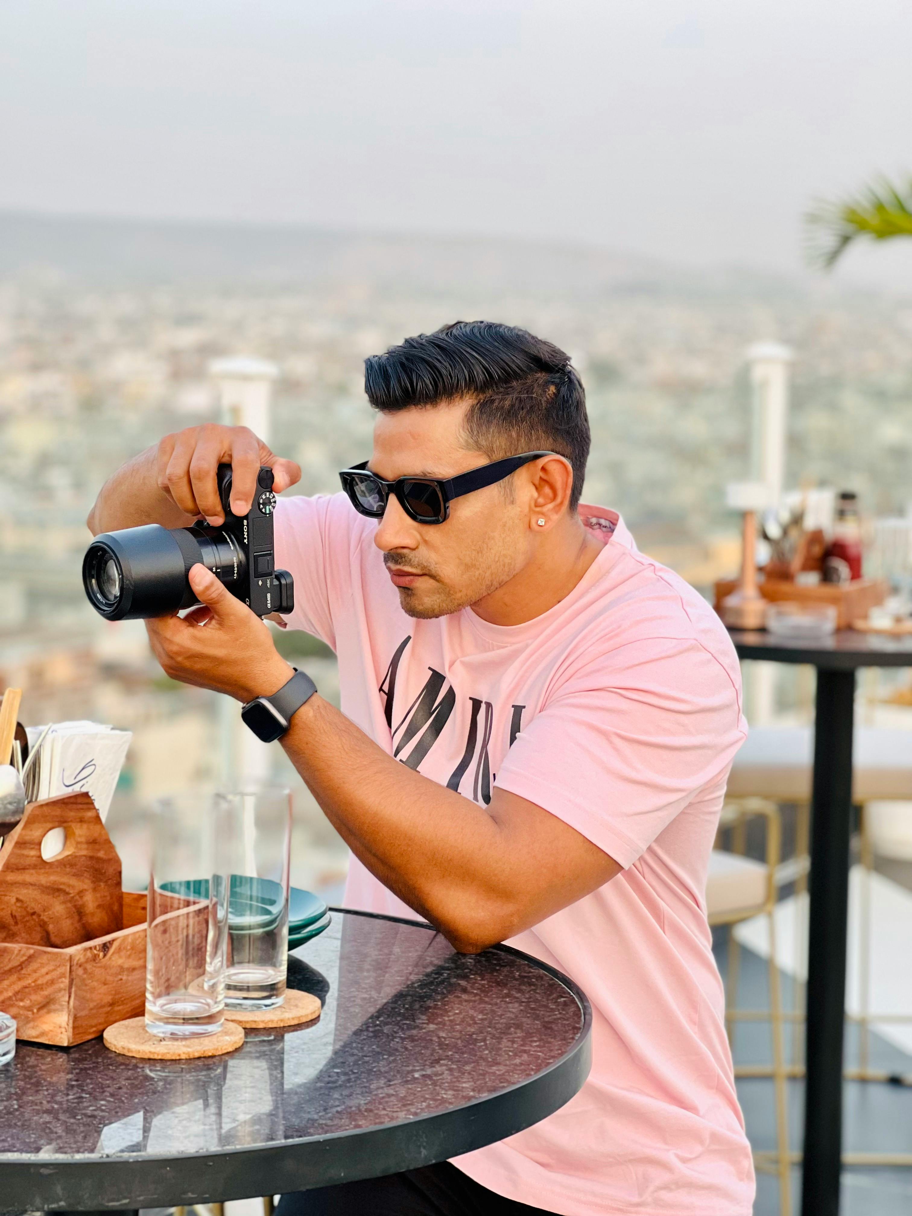 Adult man in pink shirt and sunglasses taking photos at urban rooftop cafe.
