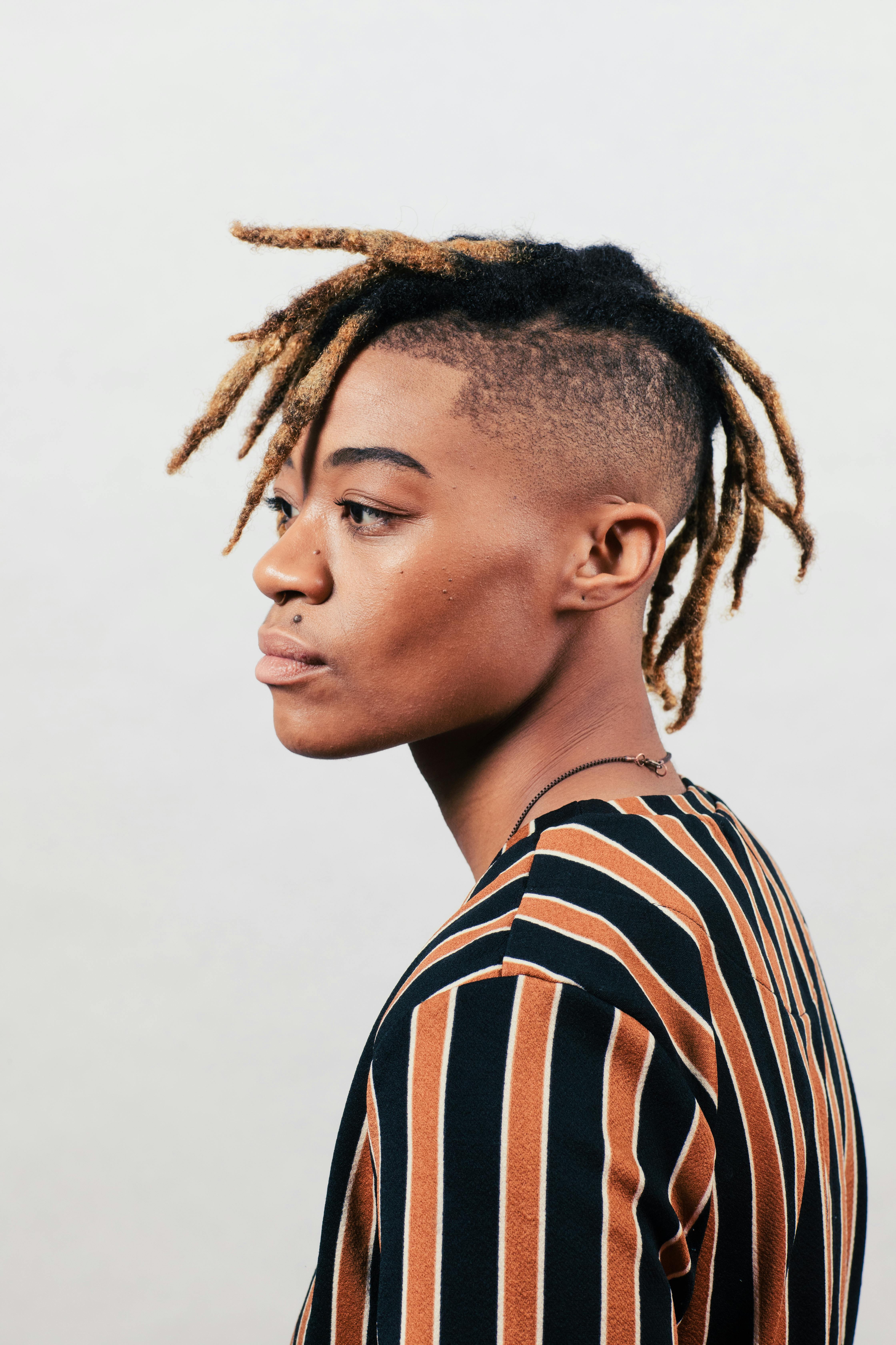 Side profile portrait of an adult with dreadlocks wearing a striped shirt against a white background.