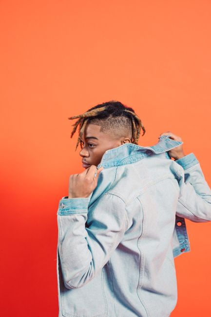 Stylish individual posing in a denim jacket against a vibrant orange studio backdrop.