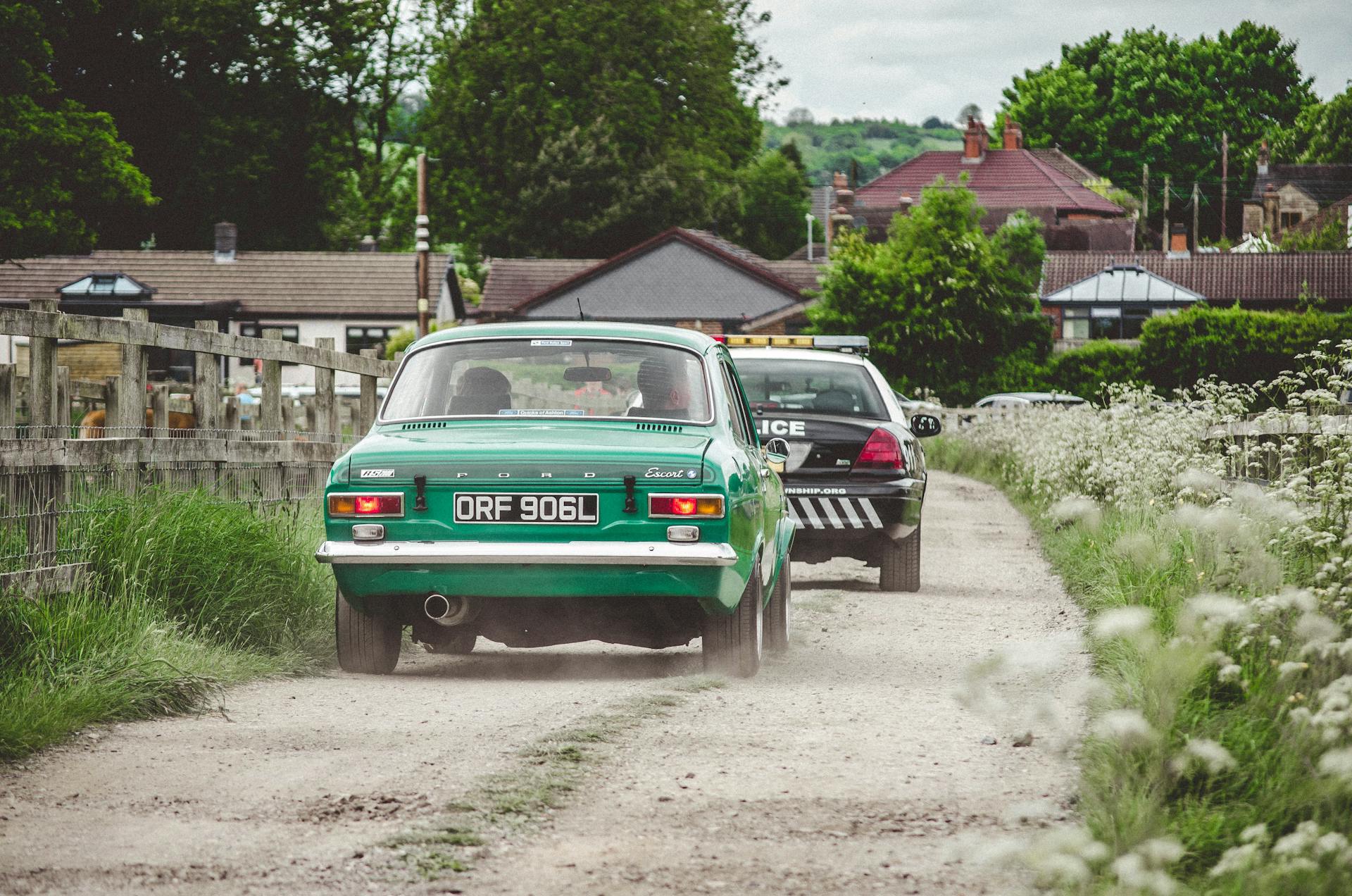 Vintage car being chased by police on a dirt road in a countryside town.