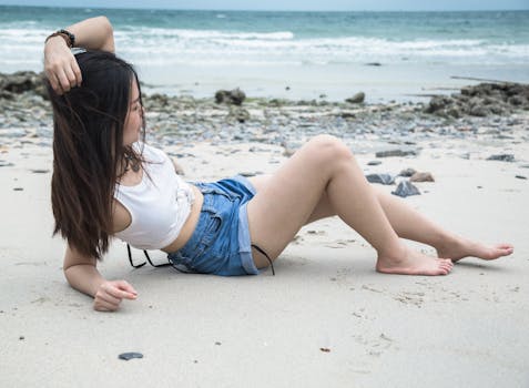 A young woman in denim shorts and a white tank top relaxes on a scenic sandy beach.