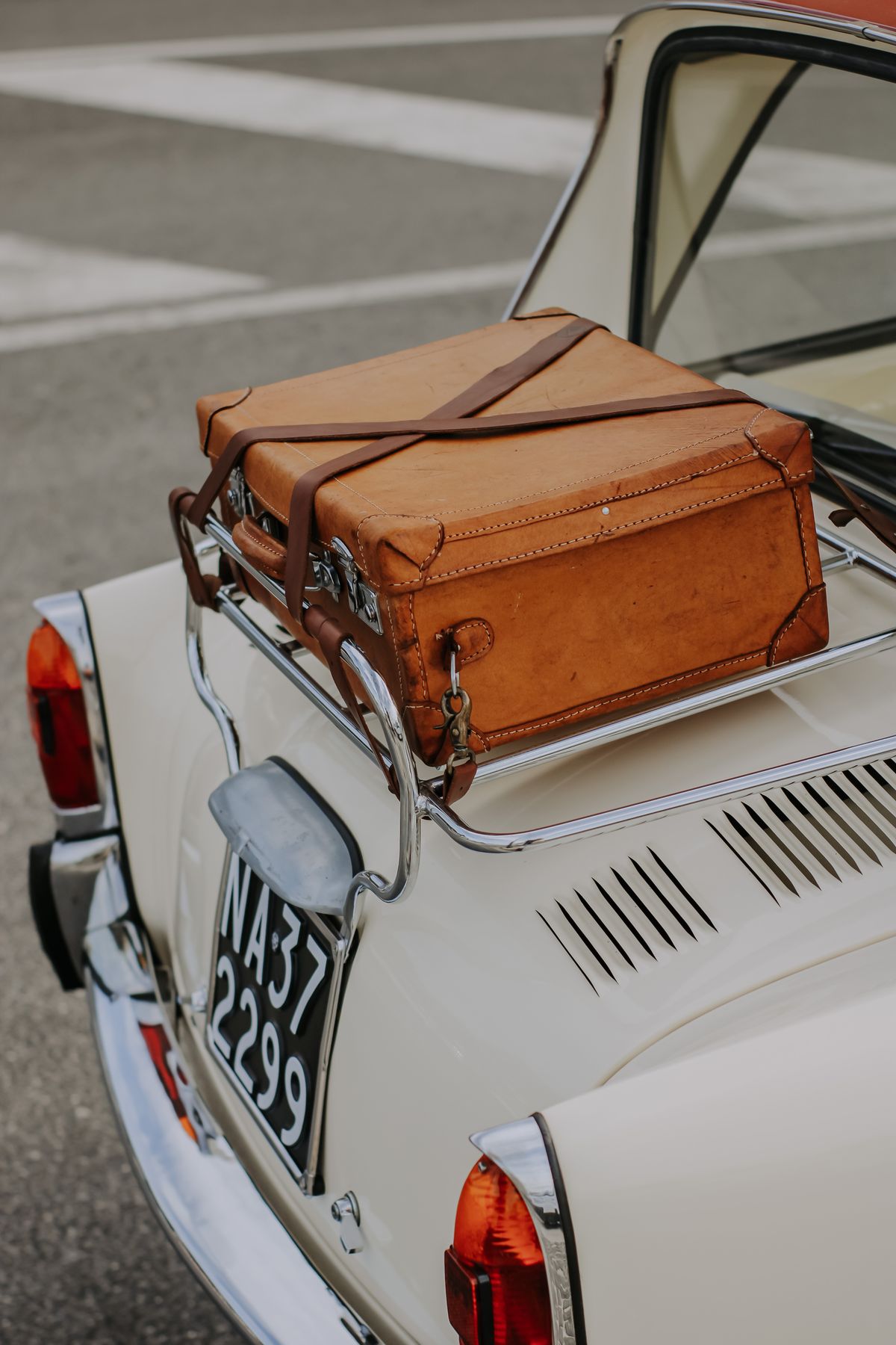 A leather suitcase strapped to the rack of a cream vintage car on an Italian street