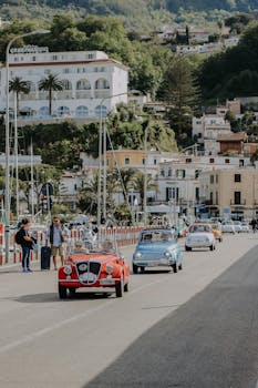 Classic vintage cars cruising along a lively coastal road with vibrant town backdrop.