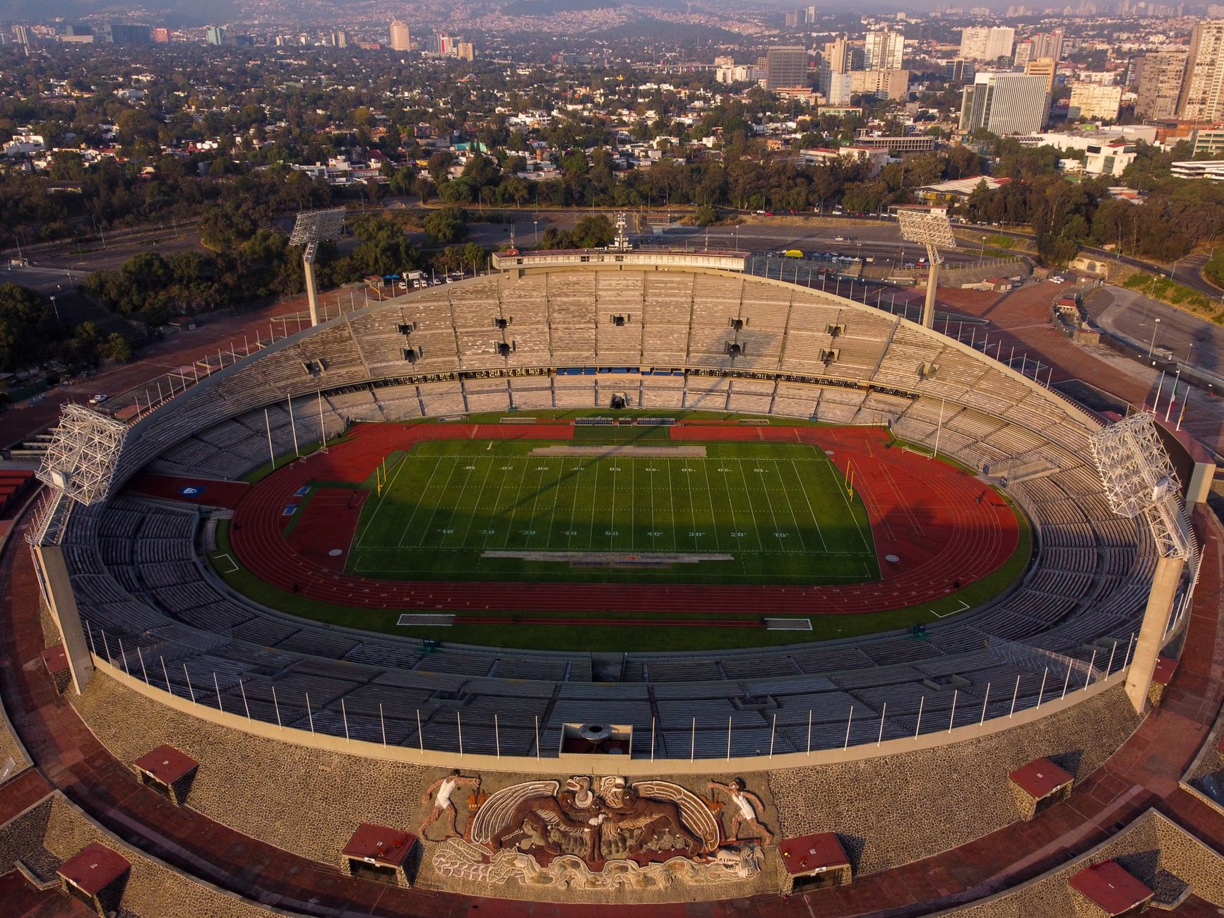 An aerial view of an empty Olympic stadium with a soccer field during daylight in an urban setting.