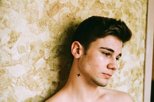 Close-up portrait of a serious young man with a tattoo, leaning against a textured wall.