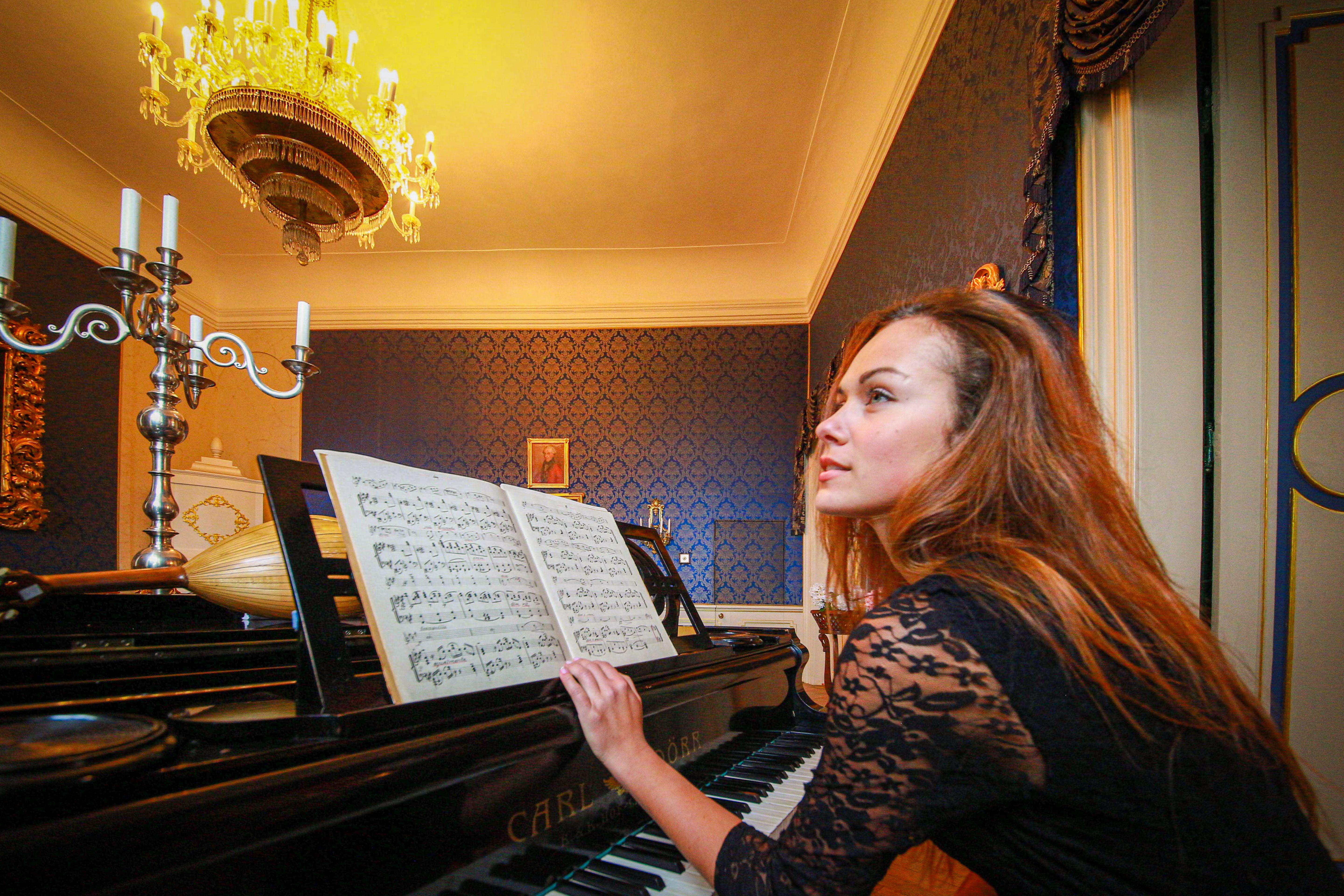 Woman plays piano in a lavish room with ornate candelabra and chandelier, creating a serene musical atmosphere.
