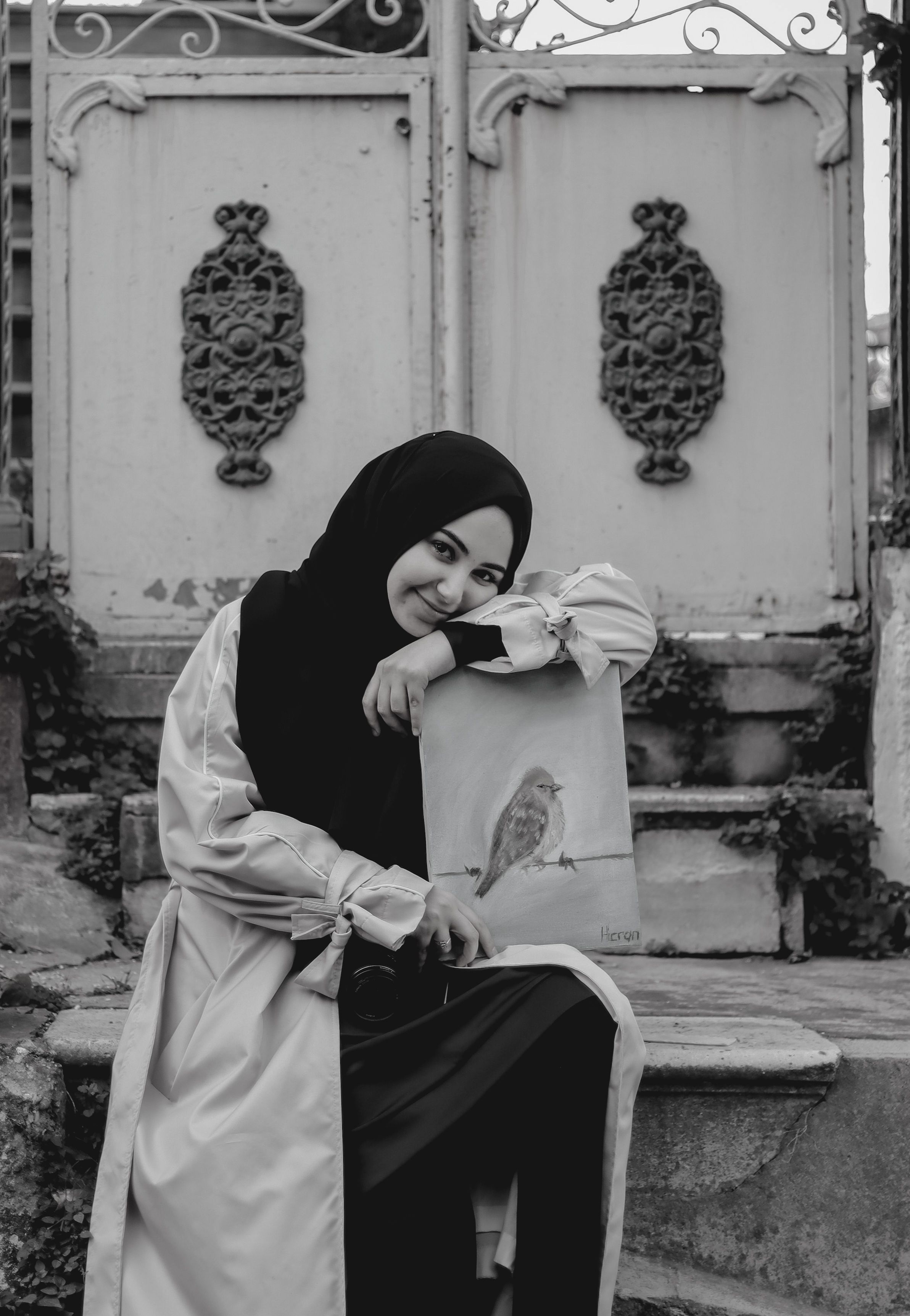 Captivating black and white portrait of a woman with a bird painting, seated in front of an ornate gate.