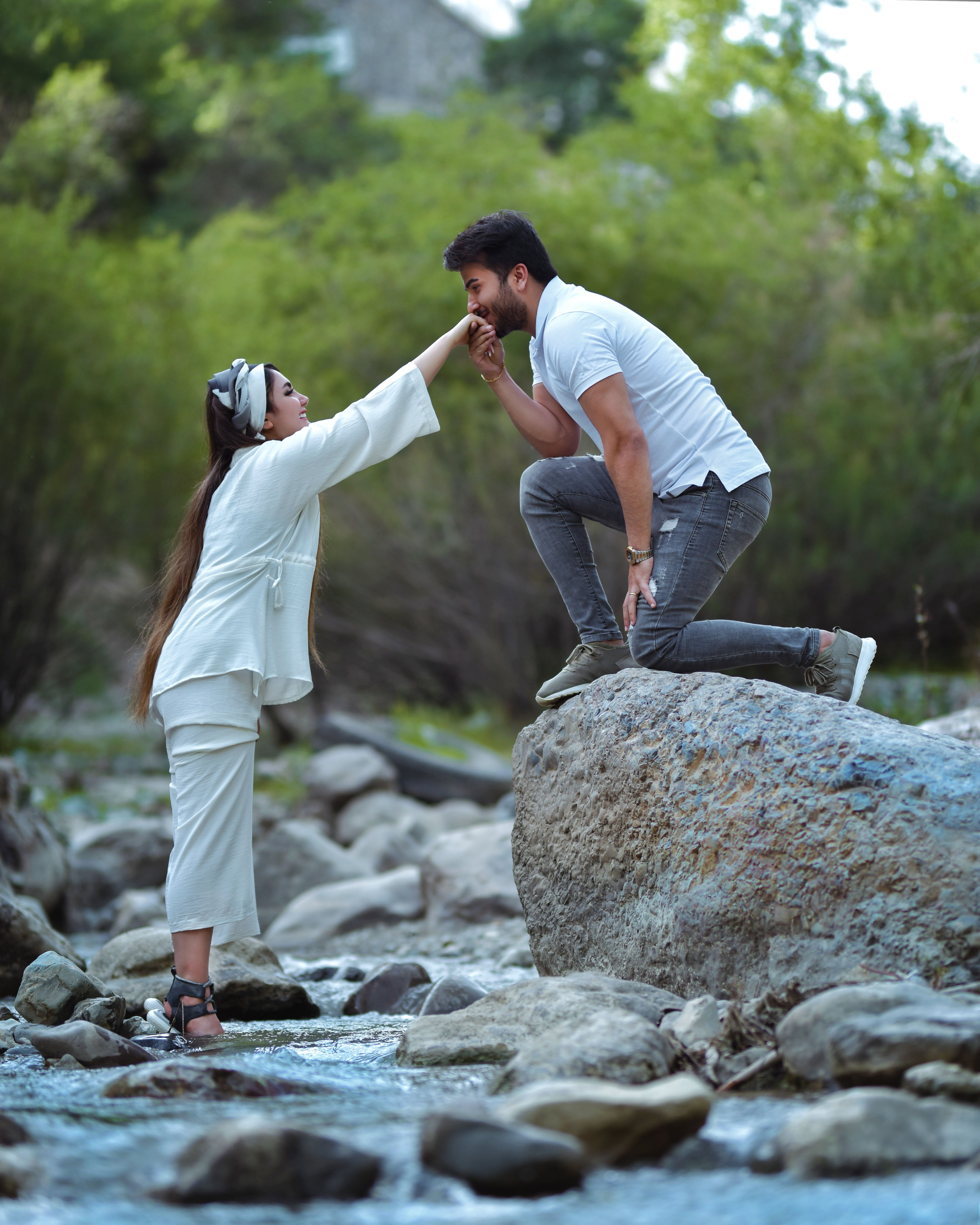 Couple sharing a romantic gesture by a rocky riverbed, surrounded by lush greenery.