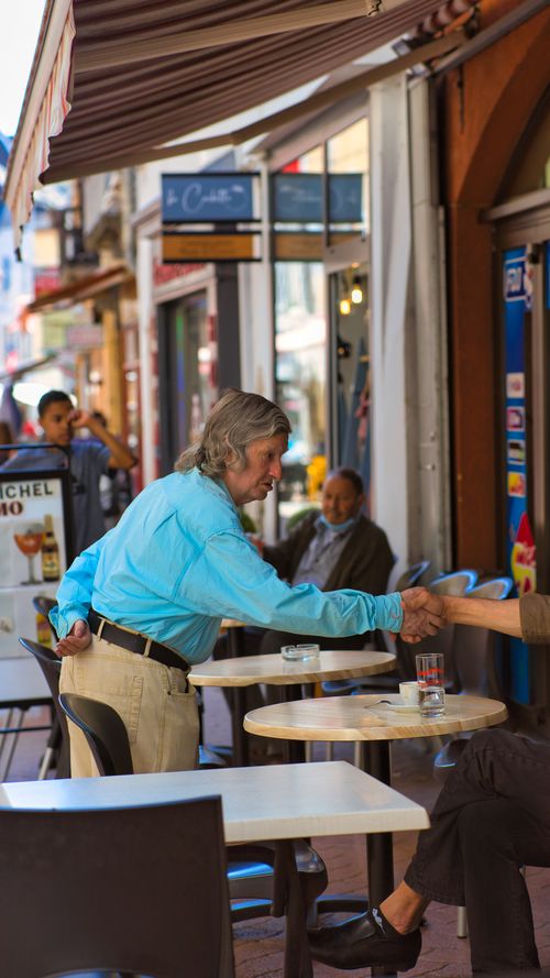 Free Two adults casually shaking hands at an outdoor café, showcasing urban street life. Stock Photo