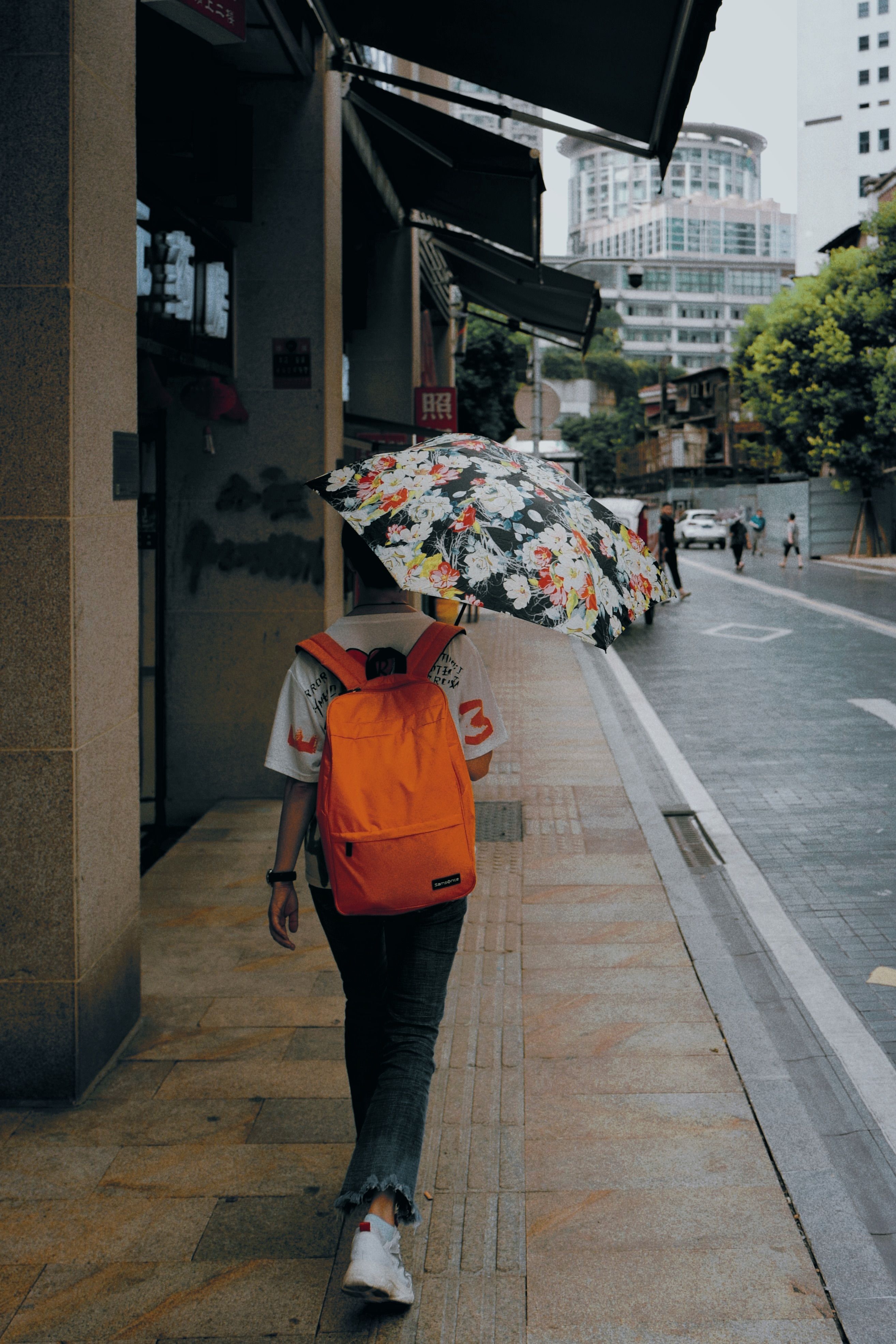 Person walking on city sidewalk with a floral umbrella and orange backpack.