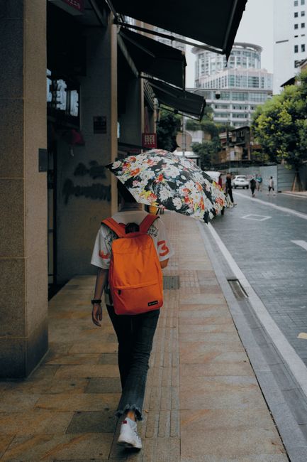 Person walking on city sidewalk with a floral umbrella and orange backpack.