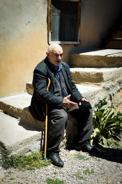 Free Senior man in a plaid blazer with a cane relaxing on sunlit concrete steps outdoors. Stock Photo