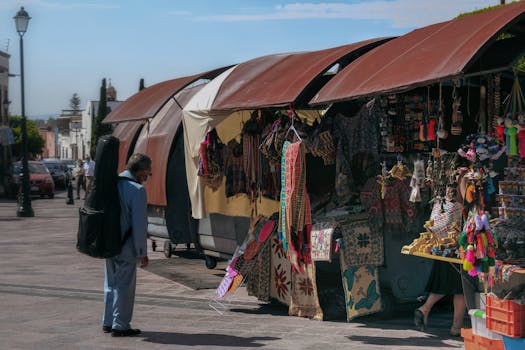 A man with a guitar case observes colorful market stalls with handcrafted goods on a sunny street.